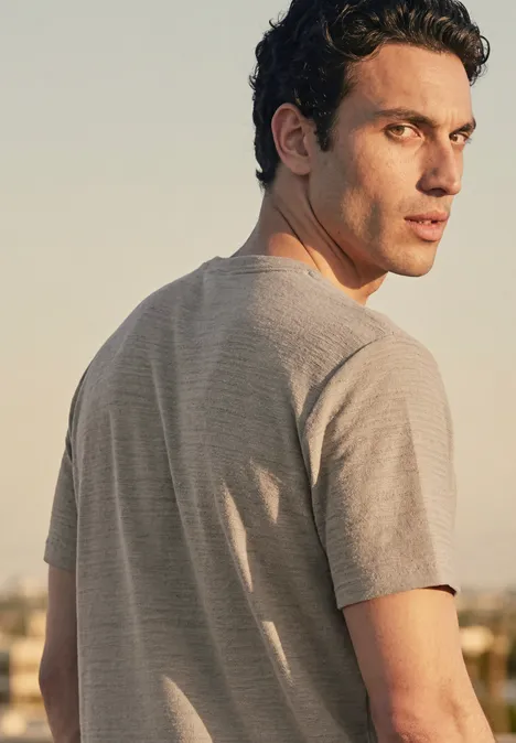 Ein Mann in grauem T-Shirt blickt über die Schulter in die Kamera, vor einer urbanen Abendkulisse.