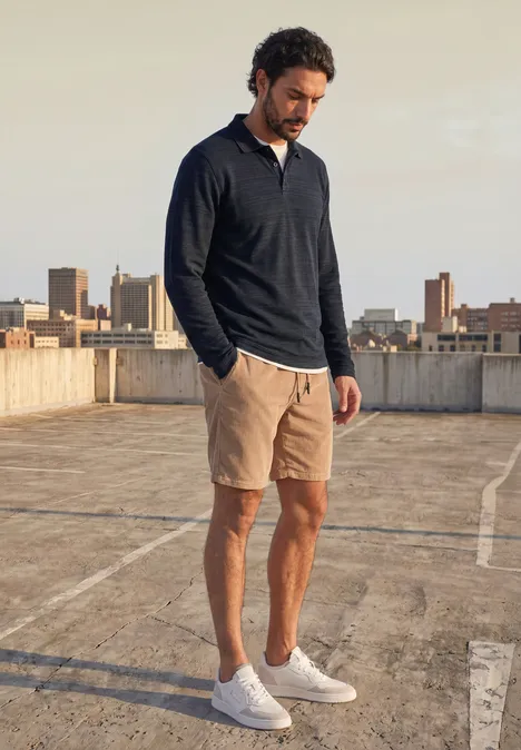 Ein Mann steht auf einem Parkdeck mit Stadtkulisse, trägt dunkelblauen Pullover, beige Shorts und weiße Sneaker.