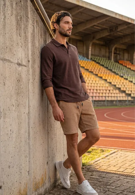Ein Mann lehnt an einer Betonmauer auf einer Sportanlage, trägt braunes Oberteil, kurze Hose und weiße Sneaker.
