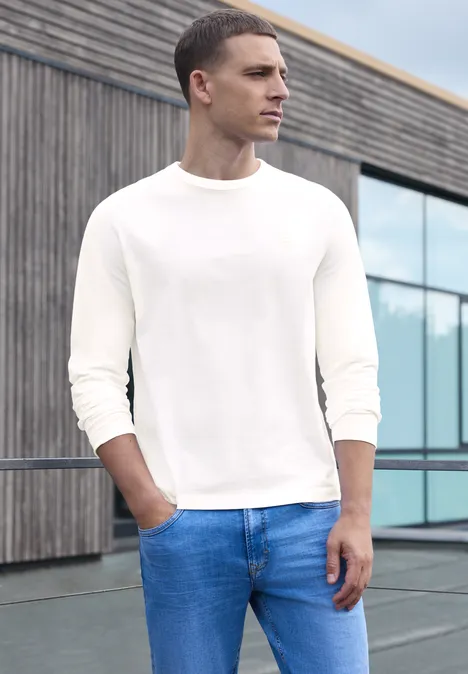 Ein Mann trägt ein weißes Langarmshirt und blaue Jeans, steht draußen vor einem modernen Gebäude.