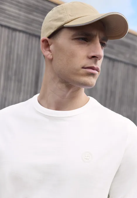 Ein Mann mit beigefarbener Cap und weißem T-Shirt vor Holzwand