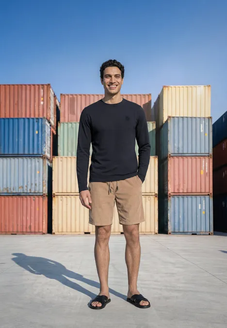 Ein lächelnder Mann steht vor bunten Frachtcontainern, trägt Langarmshirt, Shorts und Sandalen.