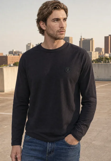 Langarm Shirt mit Rundhals in Unifarbe basic black Ein Mann in schwarzem Pullover und Jeans steht auf einem Parkdeck mit Städtesilhouette im Hintergrund.