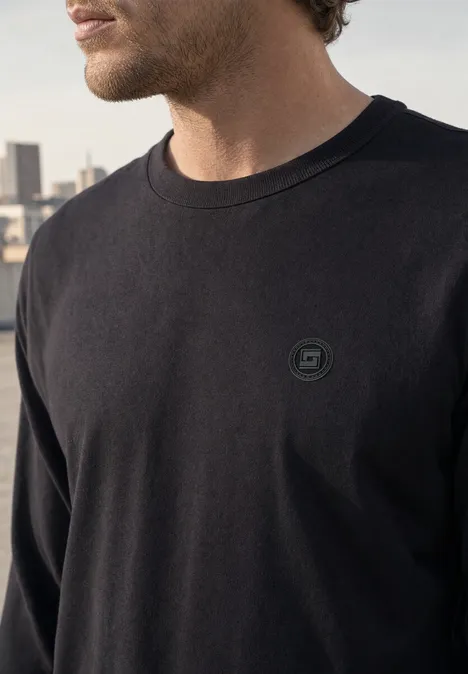 Langarm Shirt mit Rundhals in Unifarbe basic black Mann in schwarzem Langarmshirt mit kleinem Logo, davor unscharfe Stadtsilhouette.