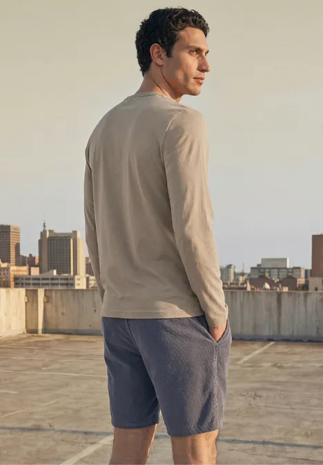 Ein Mann steht auf einem Dach und blickt über die Stadt, trägt ein beiges Langarmshirt und blaue Shorts.
