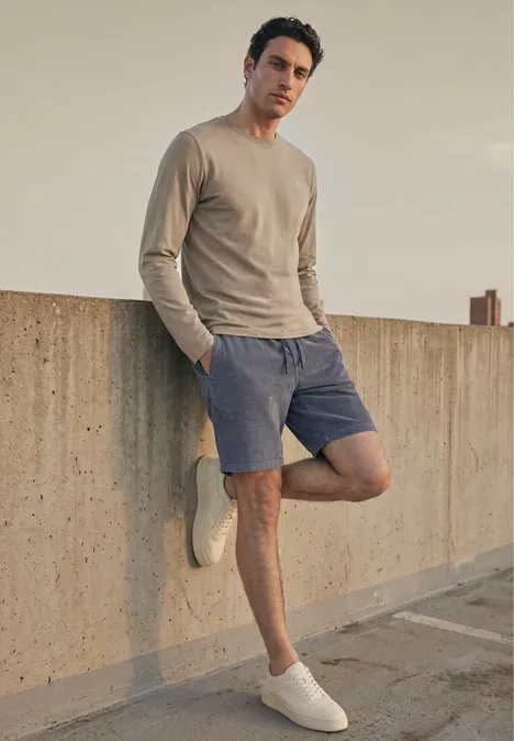 Junger Mann in beigem Pullover und blauen Shorts lehnt lässig an einer Betonmauer auf dem Dach.