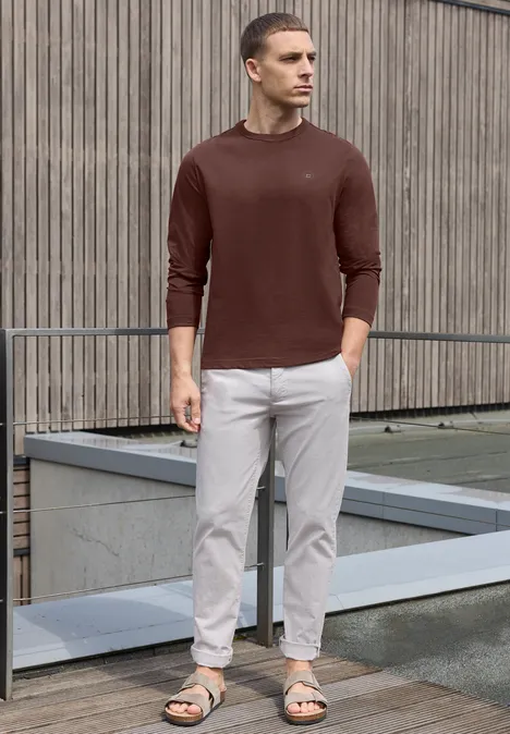 Ein Mann steht vor einer Holzfassade, trägt einen braunen Pullover, helle Chinos und Sandalen.
