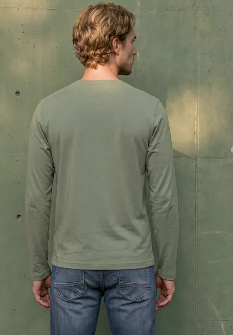 Man van achteren in een groen langarmshirt en spijkerbroek, staand voor een groene muur.