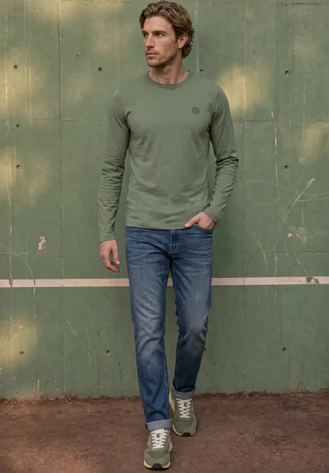 Een man in een groen shirt met lange mouwen, jeans en groene sneakers voor een groene muur op een tennisbaan.