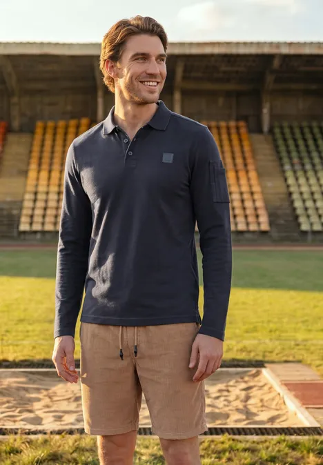Man in donkerblauw poloshirt met lange mouwen en beige short glimlacht op een atletiekbaan voor lege tribunes.