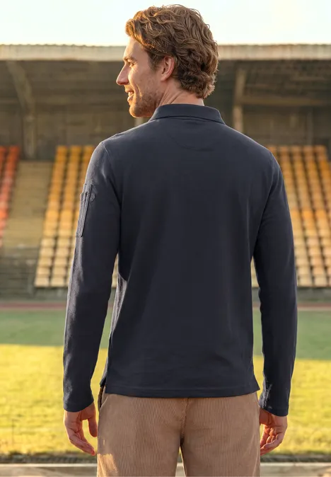 Ein Mann steht mit dem Rücken zur Kamera auf einem Stadionrasen, trägt blaues Langarmshirt und braune Hose.