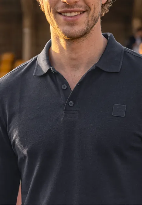 Ein lächelnder Mann trägt ein dunkelgraues Poloshirt vor unscharfem Hintergrund.
