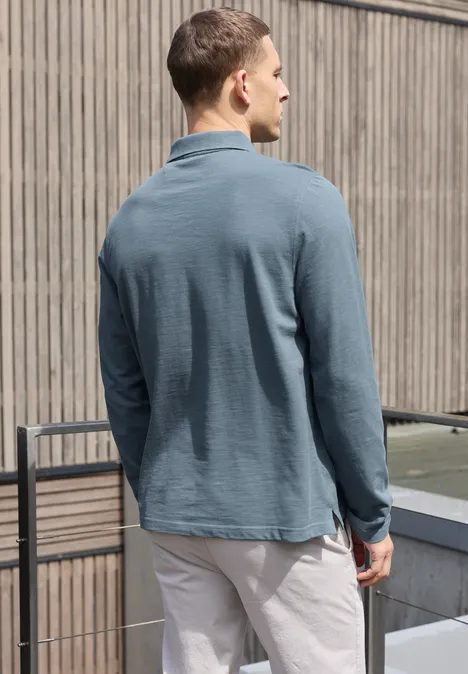 Ein Mann steht von hinten mit einem petrolfarbenen Langarm-Poloshirt und hellen Hosen auf einem Balkon.