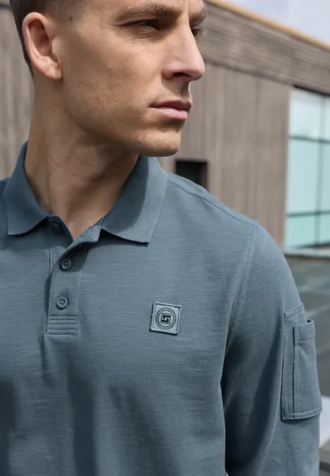 Ein Mann im Profil trägt ein graublaues Polohemd mit Brust-Patch und Ärmeltasche vor einer modernen Fassade.