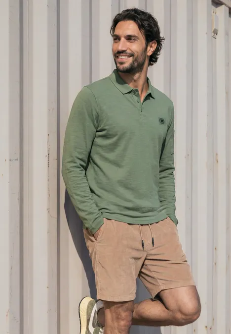 Mann lehnt lächelnd in grüner Poloshirt und beigen Shorts vor einer Metallwand.
