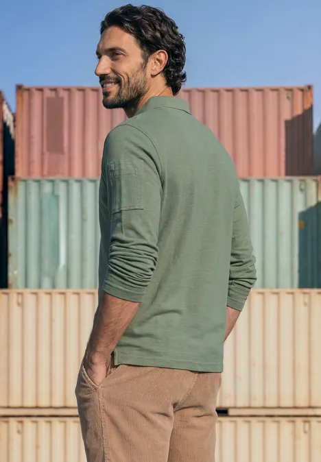 Ein bärtiger Mann in grünem Langarmshirt und brauner Hose blickt über die Schulter vor bunten Frachtcontainern.