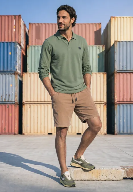 Ein Mann in grünem Poloshirt, braunen Shorts und Sneakern steht vor bunten Frachtcontainern.