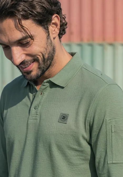 Ein junger Mann in grünem Poloshirt lächelt, den Blick nach unten gerichtet vor pastellfarbenem Hintergrund.