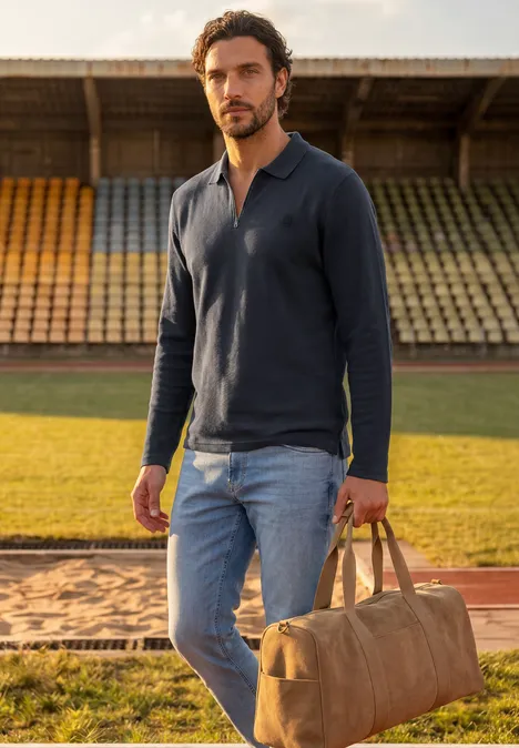 Ein Mann in Jeans und dunkelblauem Hemd trägt eine Reisetasche auf einer Sportanlage.