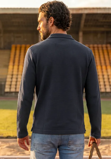 Ein Mann trägt einen dunklen Pullover und hält einen Ball auf einer Stadiontribüne.