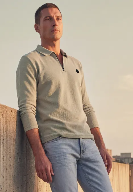 Ein Mann lehnt an einer Mauer, trägt helles Poloshirt und Jeans und blickt im Abendlicht in die Ferne.