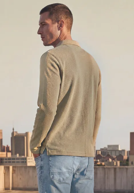 Mann im hellen Pullover und Jeans, der auf einem Dach mit Stadtkulisse steht.