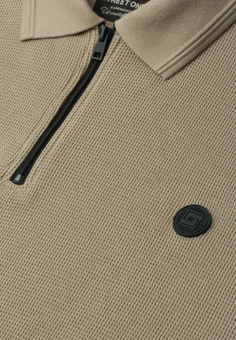 Beige Strick-Polo mit Reißverschluss und rundem schwarzem Logo-Emblem.