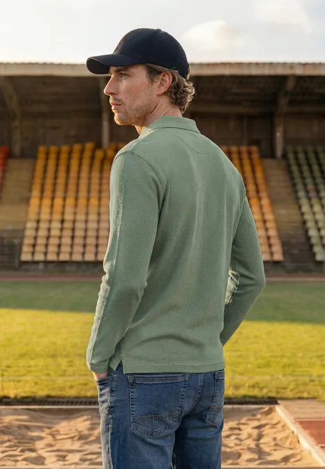 Ein Mann im grünen Langarm-Poloshirt und Cap steht seitlich vor leeren Tribünen.