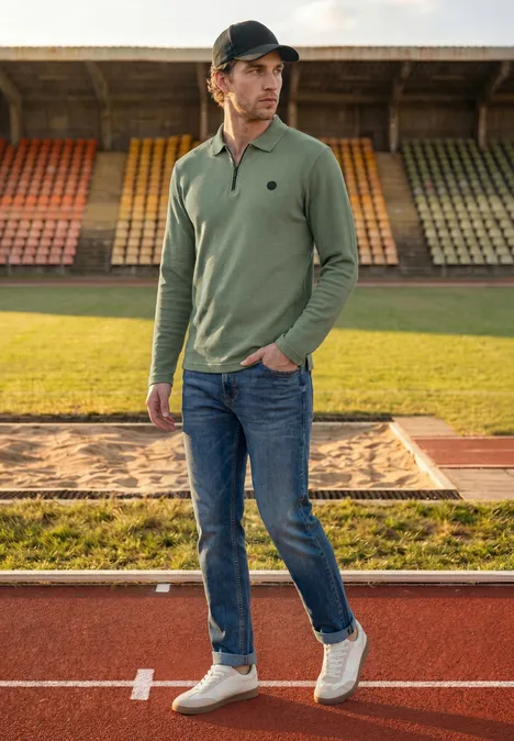 Ein junger Mann mit grünem Langarm-Poloshirt, Jeans und Cap steht auf der Laufbahn vor Tribünen.