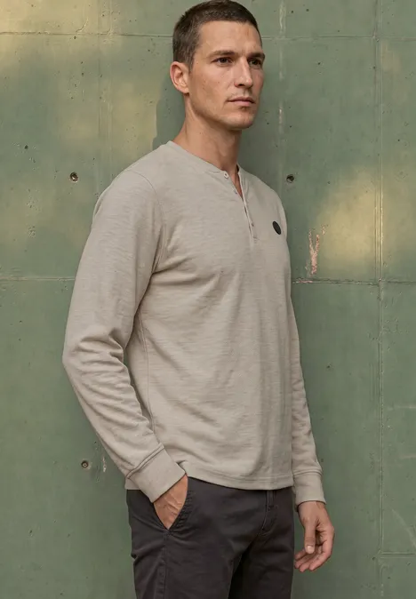 Ein Mann trägt ein graues Langarm-Henley-Shirt, steht seitlich mit Händen in den Hosentaschen vor einer grünen Wand.