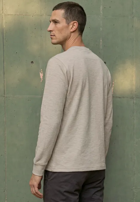 Seitliche Ansicht eines Mannes in grauem Langarmshirt und dunkler Hose vor grünem Hintergrund.
