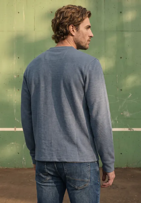 Ein Mann steht von hinten in Jeans und langärmeligem Shirt vor einer grünen Wand.