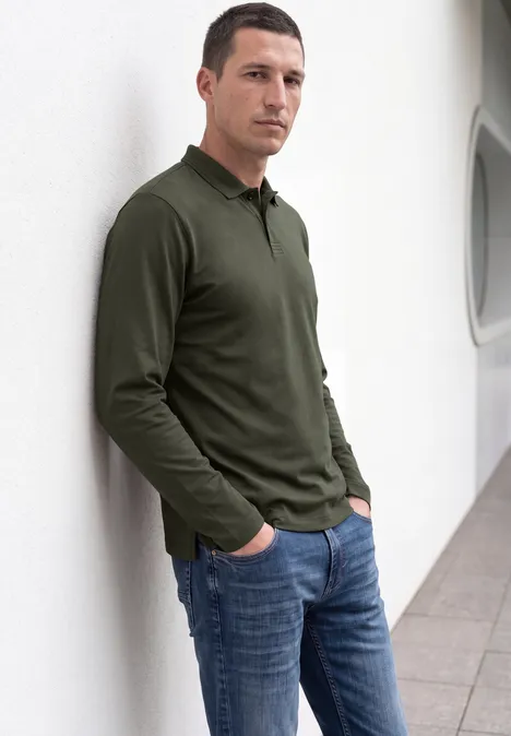Ein Mann lehnt lässig in einem weißen Flur an einer Wand, trägt olivgrünes Langarm-Poloshirt und blaue Jeans.