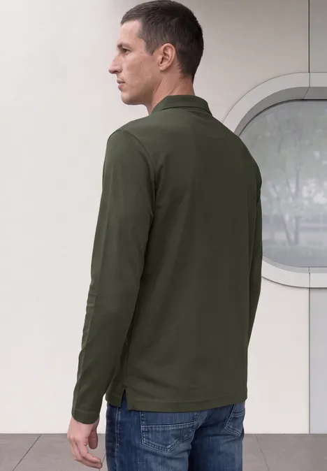 Mann von hinten in dunkelgrünem Langarm-Polo und Jeans vor hellem Hintergrund mit rundem Fenster.