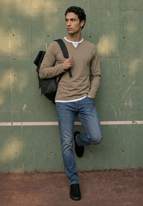 Henleyshirt im 2-in-1-Look antique beige Ein Mann lehnt an einer grünen Wand, trägt Jeans, Pulli und Rucksack, schaut in die Ferne.