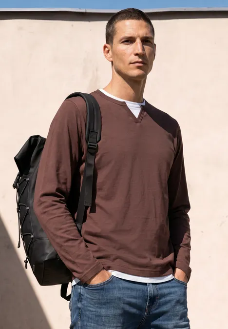 Mann in braunem Langarmshirt, weißem Innenhemd und mit Rucksack steht vor einer hellen Wand.