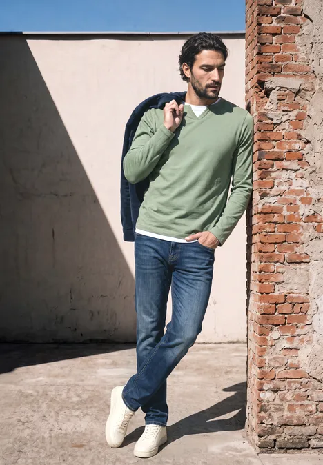 Henleyshirt im 2-in-1-Look willow green Mann lehnt in sonnigem Innenhof an Backsteinwand, trägt Jeans, grünen Pullover, Jacke über der Schulter und schaut weg.