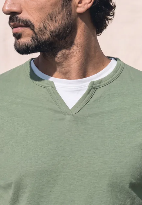 Henleyshirt im 2-in-1-Look willow green Ein Mann mit kurzem schwarzen Bart trägt einen hellgrünen Pullover über weißem T-Shirt und blickt seitlich.