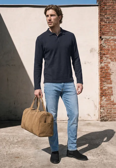 Langarm Poloshirt in Unifarbe dark ink blue Ein Mann in blauem Poloshirt, hellen Jeans und schwarzen Schuhen trägt eine sandfarbene Reisetasche vor einer Wand.