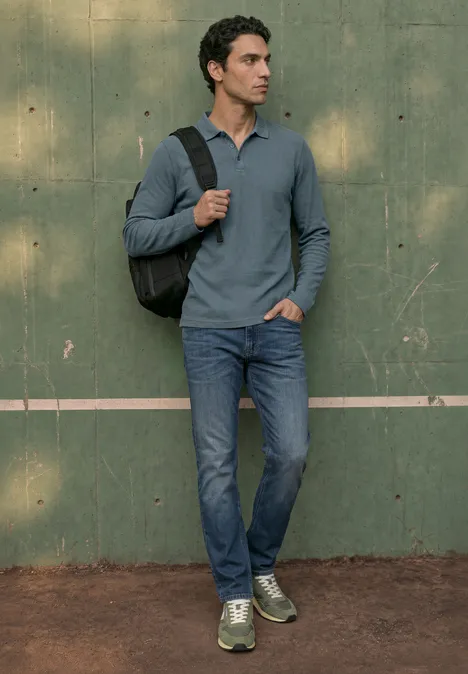 Langarm Poloshirt in Unifarbe vintage blue Ein Mann in Jeans, grauem Henley-Shirt und grünen Sneakern steht lässig mit Rucksack vor einer grünen Wand.