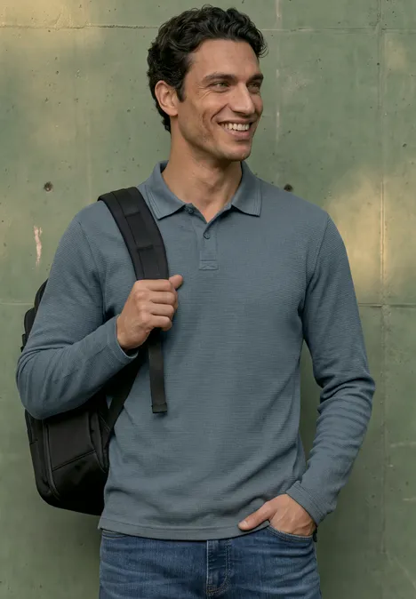 Langarm Poloshirt in Unifarbe vintage blue Mann mit Rucksack, lächelnd, vor grüner Wand.