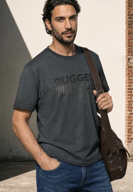 Ein Mann in dunkelgrauem T-Shirt mit Aufschrift, Jeans und Umhängetasche lehnt an einer Wand.