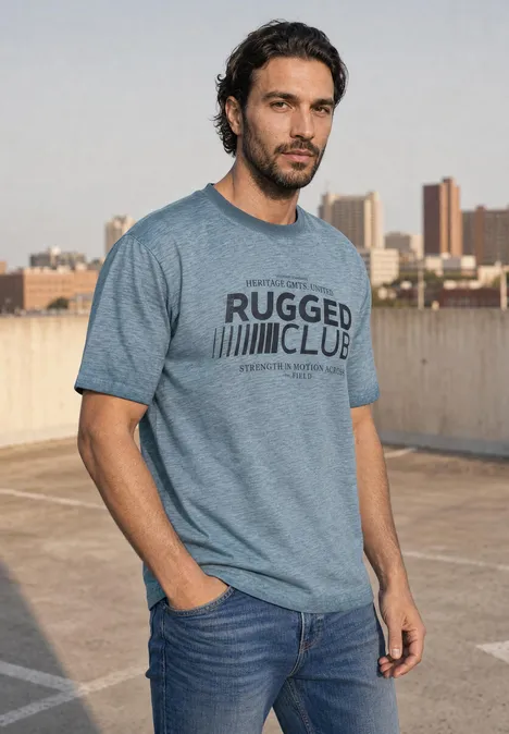 Ein Mann in Jeans und grauem T-Shirt steht auf einem Parkdeck mit Stadtkulisse im Hintergrund.
