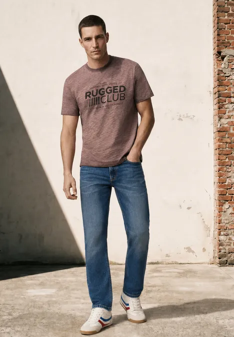 Mann in braunem T-Shirt und Jeans lehnt lässig an einer Wand
