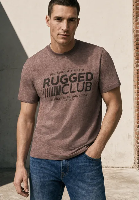 Ein Mann steht vor einer Wand, trägt ein braunes T-Shirt mit 'RUGGED CLUB'-Aufdruck und blaue Jeans.
