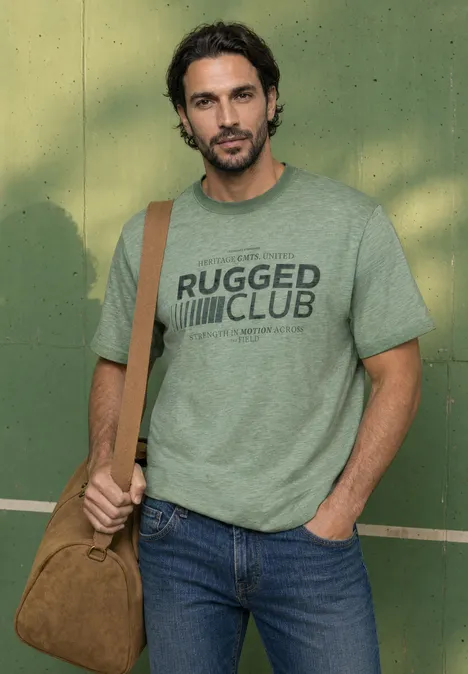 Mann mit grünem 'RUGGED CLUB'-T-Shirt, Jeans und brauner Umhängetasche steht vor grüner Wand.