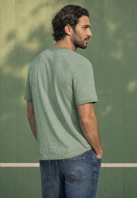 Ein Mann mit braunem Haar trägt ein grünes T-Shirt und Jeans vor einer olivgrünen Wand.
