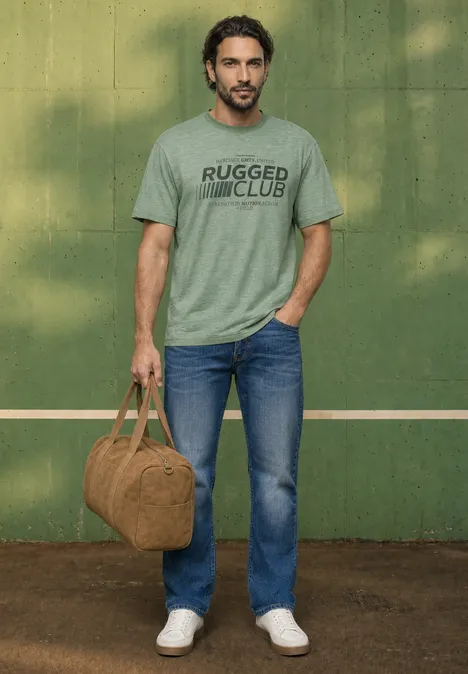 Ein Mann in Jeans und grauem T-Shirt hält eine braune Reisetasche vor einer grünen Wand.