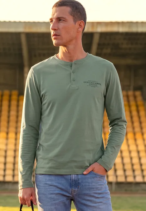 Angerautes Henleyshirt mit Printdetail willow green Ein Mann mit grünem Henley-Shirt und Jeans steht in einem Stadion, hält ein Smartphone in der Hand.