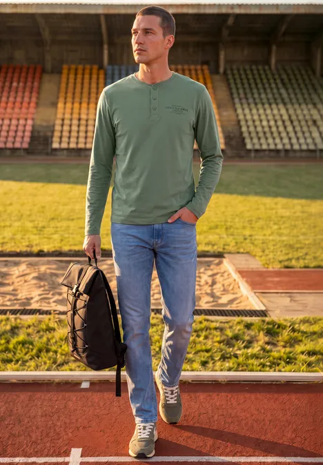 Angerautes Henleyshirt mit Printdetail willow green Ein Mann in grüner Langarmshirt und Jeans trägt einen schwarzen Rucksack auf einer Laufbahn.